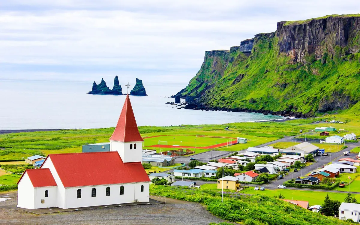 A white church with a red roof standing above the village, with green slopes and sea stacks beyond
