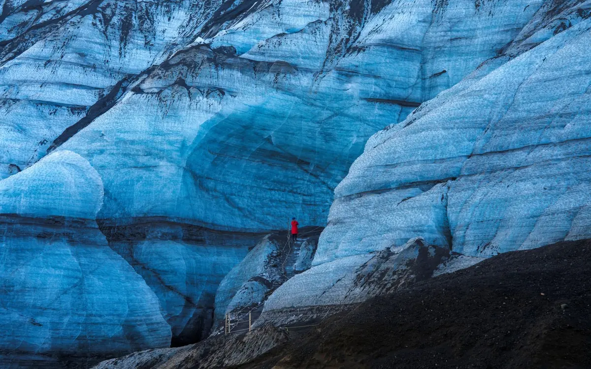 A wall of layered blue-grey glacier ice with a single tiny figure at the foot of it for scale