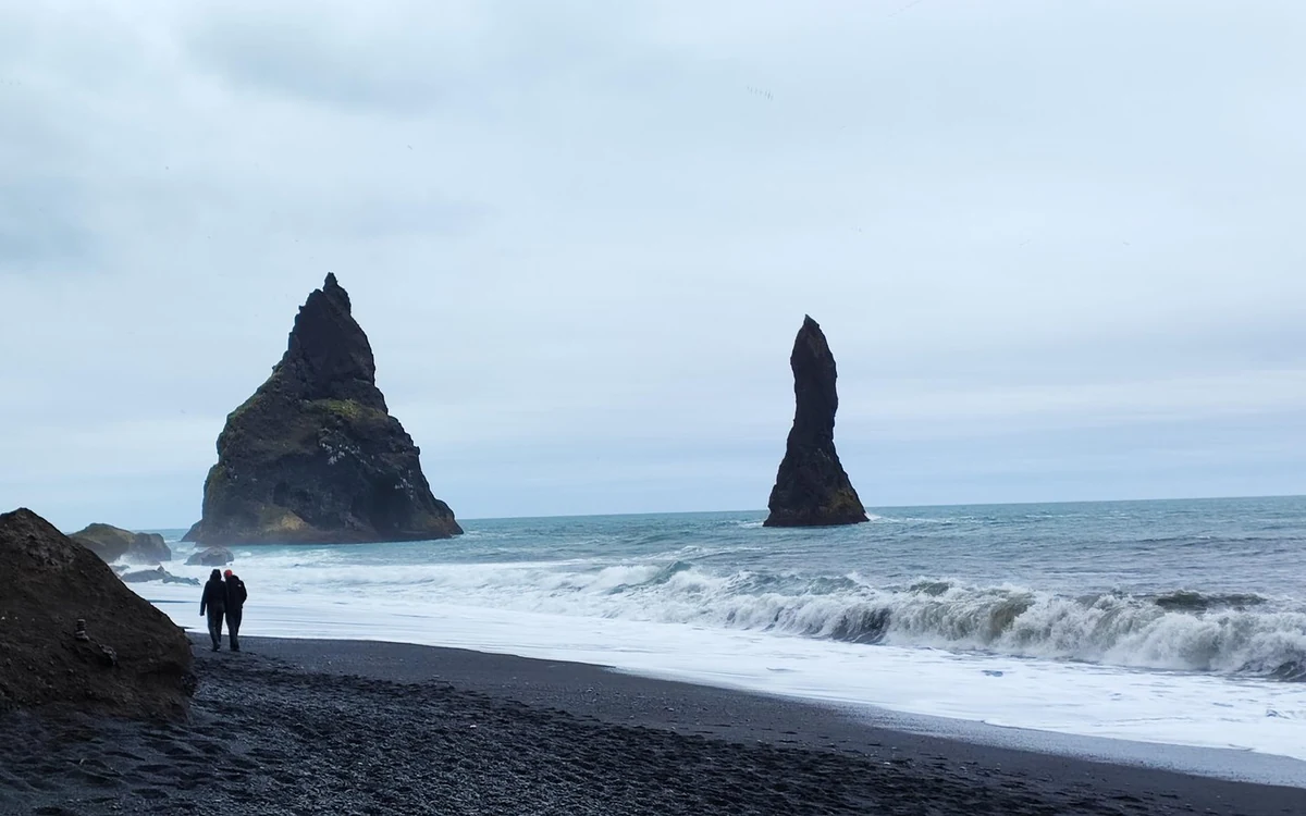 Two people walking a black sand beach with the Reynisdrangar sea stacks standing offshore