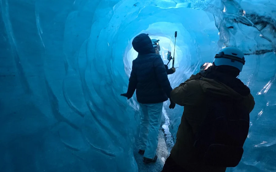 Two walkers with head torches lighting the dark blue wall of an ice tunnel