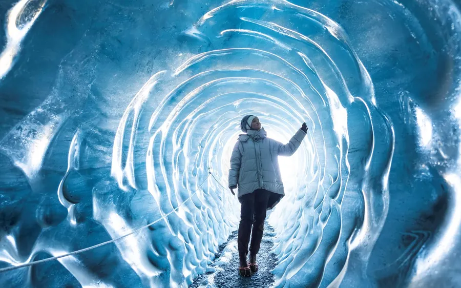 A visitor standing with arms wide in a round tunnel of ribbed blue ice