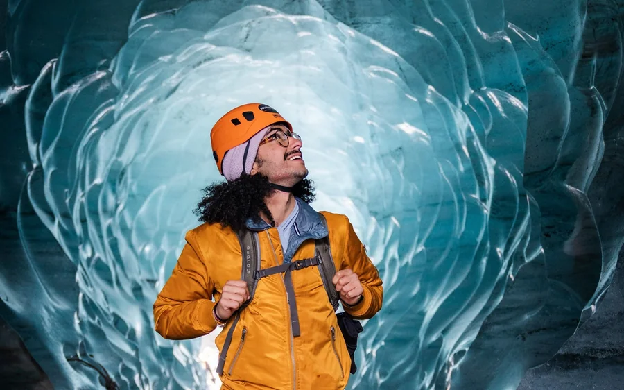 A guide in an orange jacket and orange helmet looking up inside a turquoise ice cave