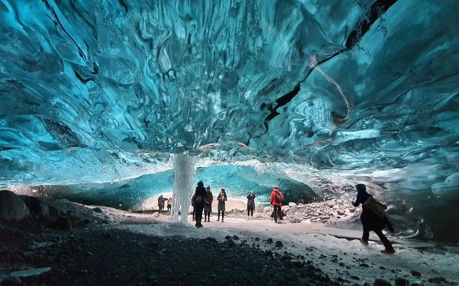 Several people walking through a large turquoise ice cavern with a gravel floor