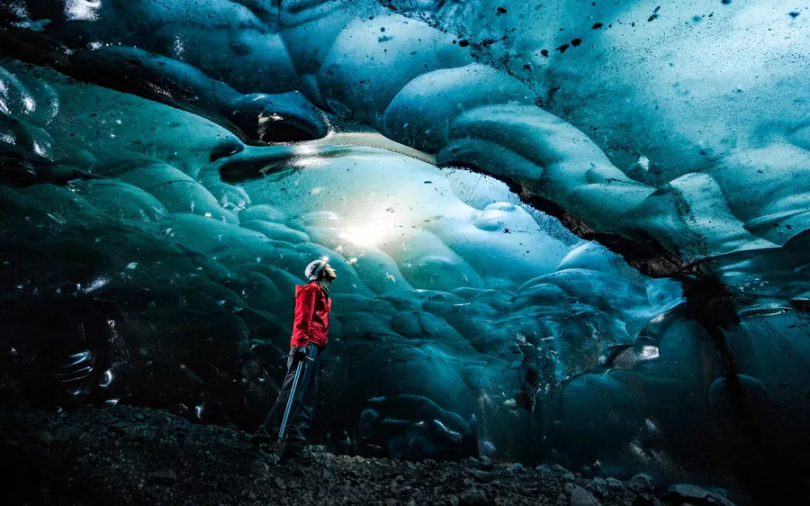 A walker in a red jacket deep inside a dark blue ice cave