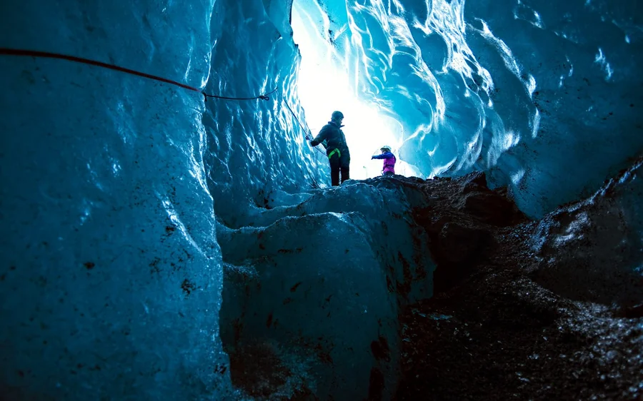 A silhouette high in a bright blue ice tunnel with a second walker below